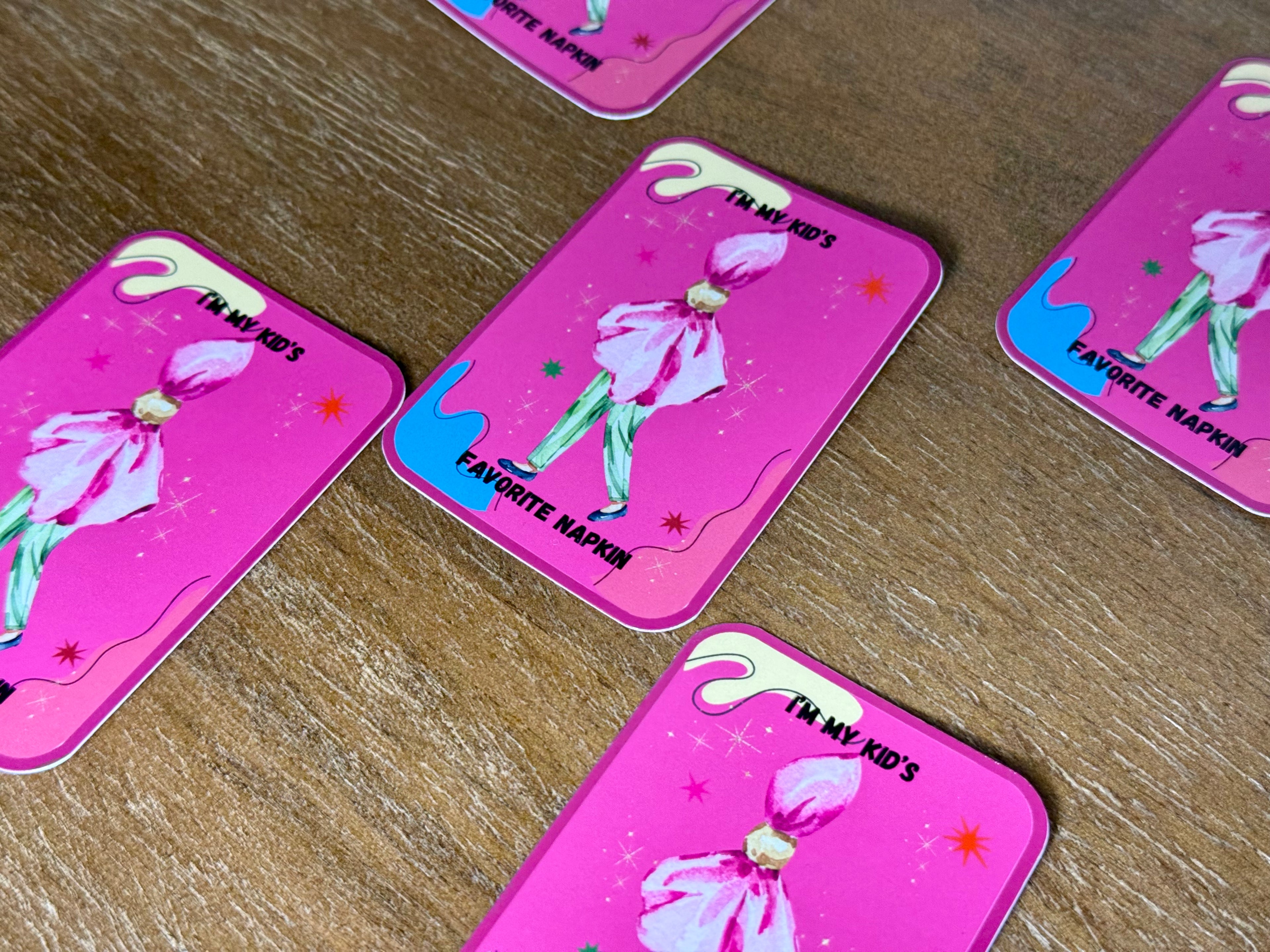 Pink rectangular cards with rounded corners arranged on a wooden surface. Each card shows a stylized figure in a pink dress, small star accents, and text including 'I'M MY KID'S' and 'FAVORITE NAPKIN.