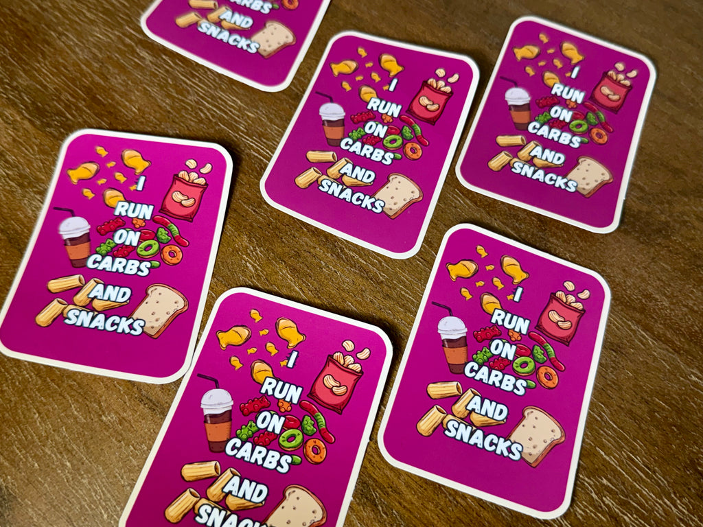 Five pink rectangular cards with rounded corners arranged on a brown wooden surface. Each card features the text 'I RUN ON CARBS AND SNACKS' in white, surrounded by cartoon illustrations of food items like chips, pizza, candy, and toast.