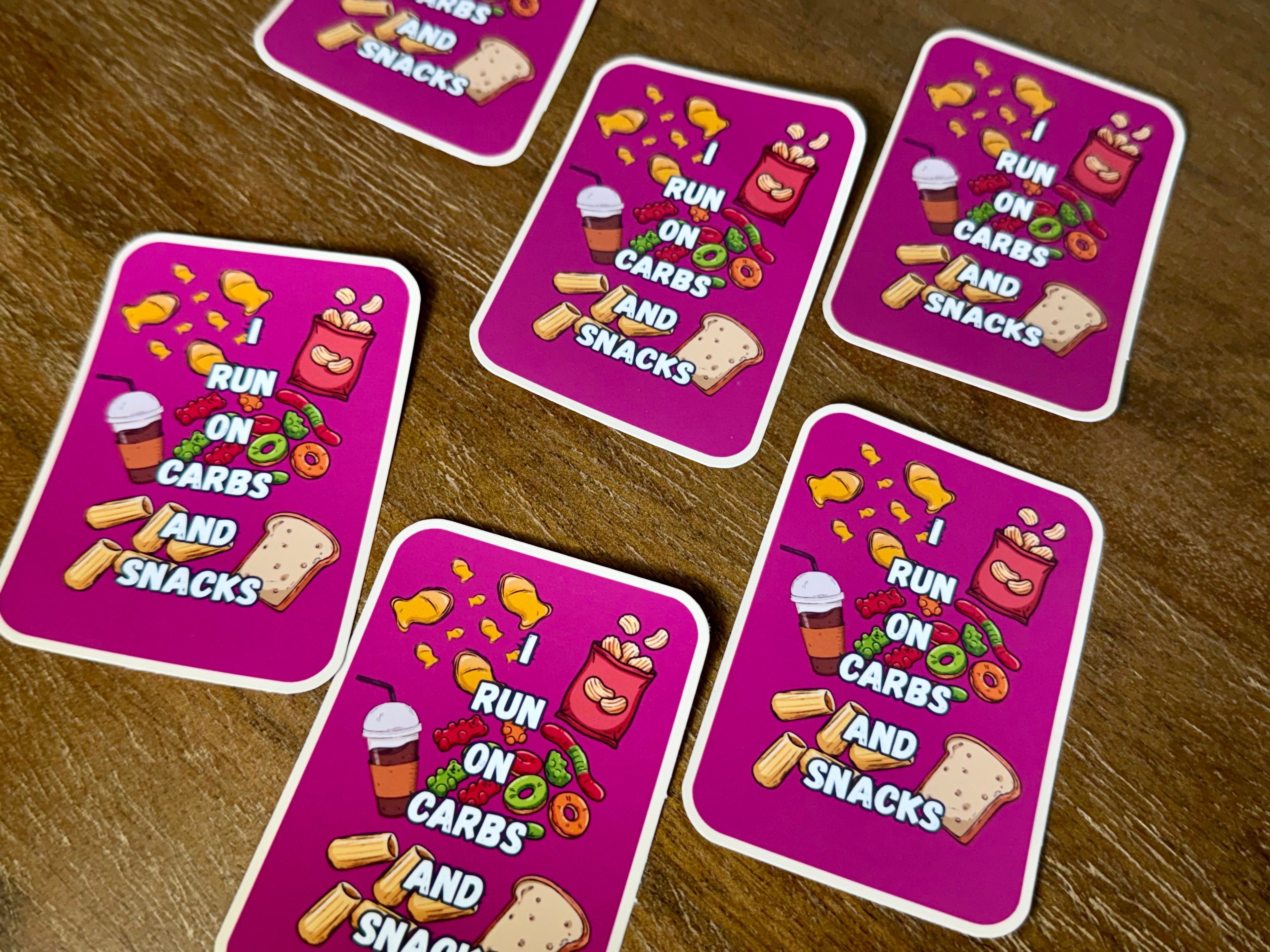 Five pink rectangular cards with rounded corners arranged on a brown wooden surface. Each card features the text 'I RUN ON CARBS AND SNACKS' in white, surrounded by cartoon illustrations of food items like chips, pizza, candy, and toast.