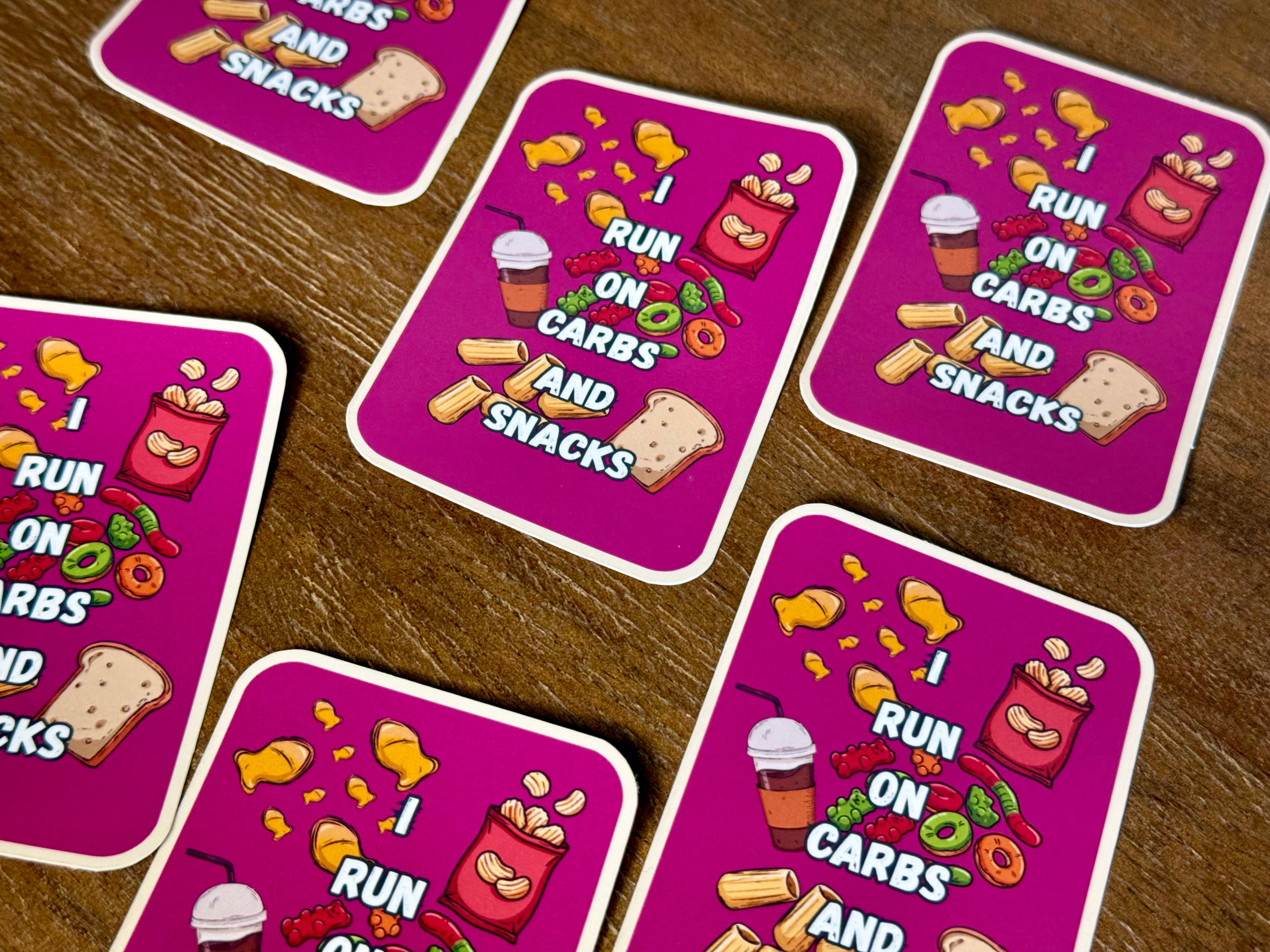 Several rounded rectangular stickers with white borders are arranged on a brown wooden surface. Each sticker has a bright pink background and cartoon-style snack illustrations, including chips, candy corn, a drink, bread, and a red bag.