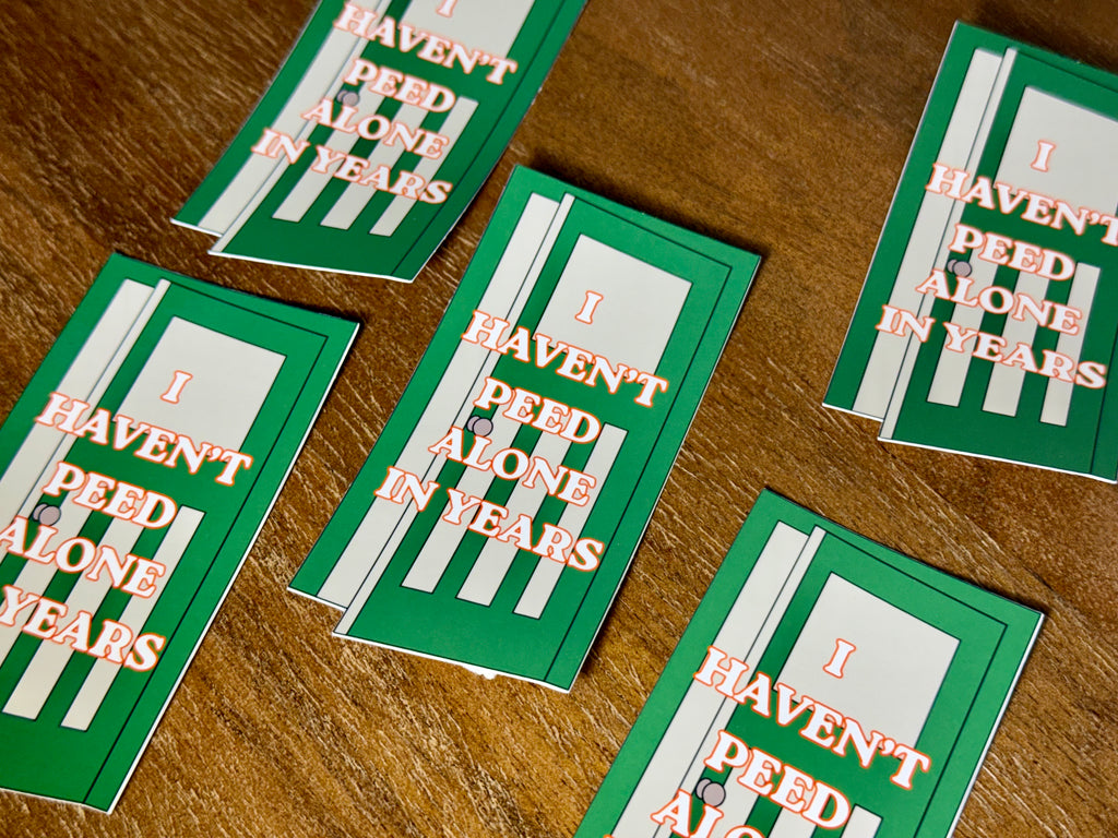 Several green and white stickers with the phrase 'I HAVEN'T PEED ALONE IN YEARS' in orange lettering, arranged on a wooden tabletop.