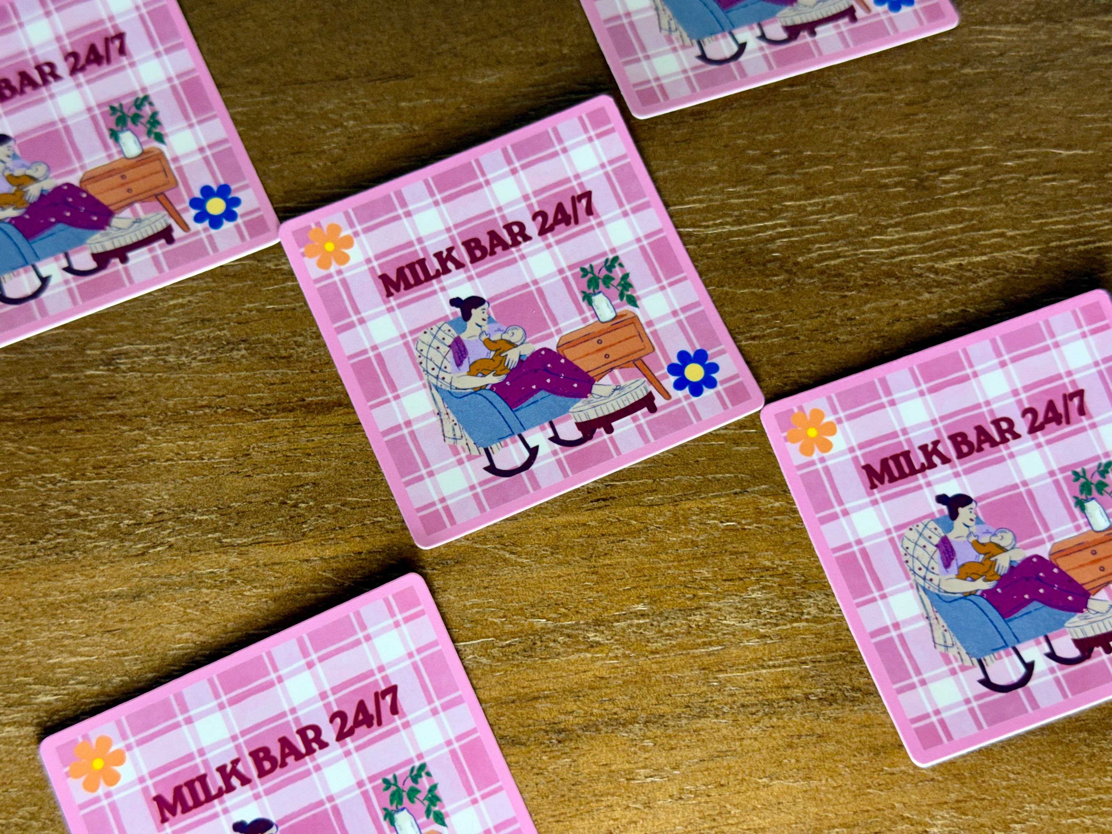 Square coasters with a pink plaid pattern and an illustration of a woman in a rocking chair, with the text 'MILK BAR 24/7' on a wooden tabletop.