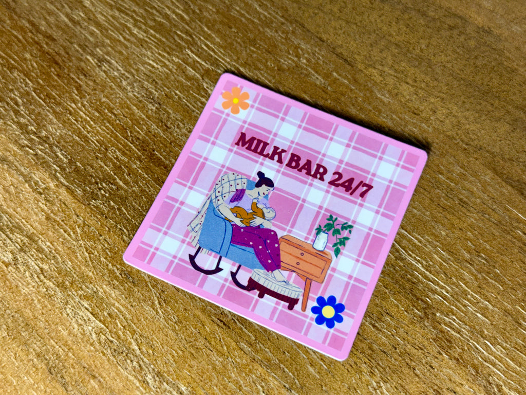 Pink "Milk Bar 24/7" sticker featuring a cozy illustration of a breastfeeding mom sitting in a chair with her baby on a plaid background.