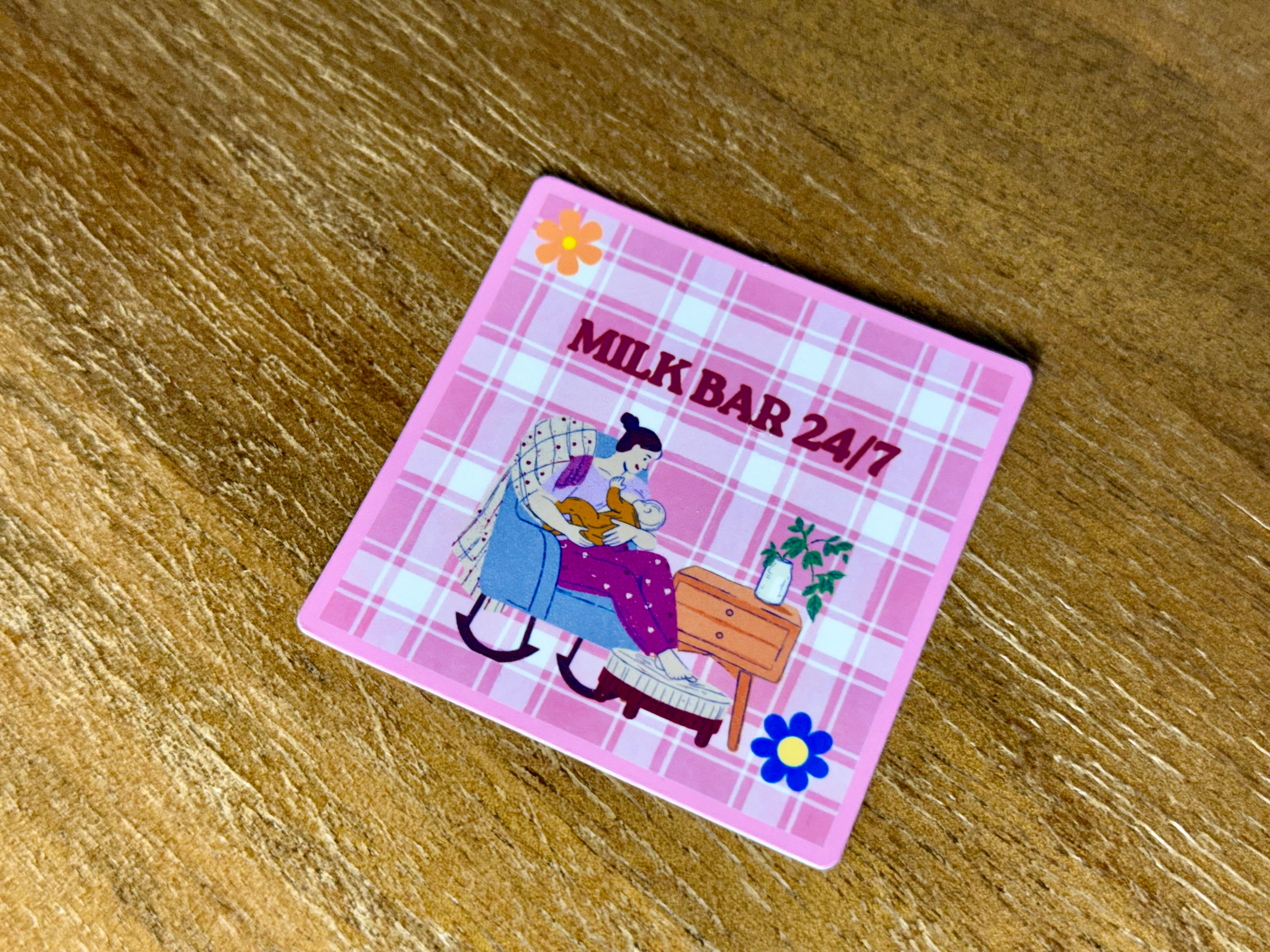 Pink "Milk Bar 24/7" sticker featuring a cozy illustration of a breastfeeding mom sitting in a chair with her baby on a plaid background.