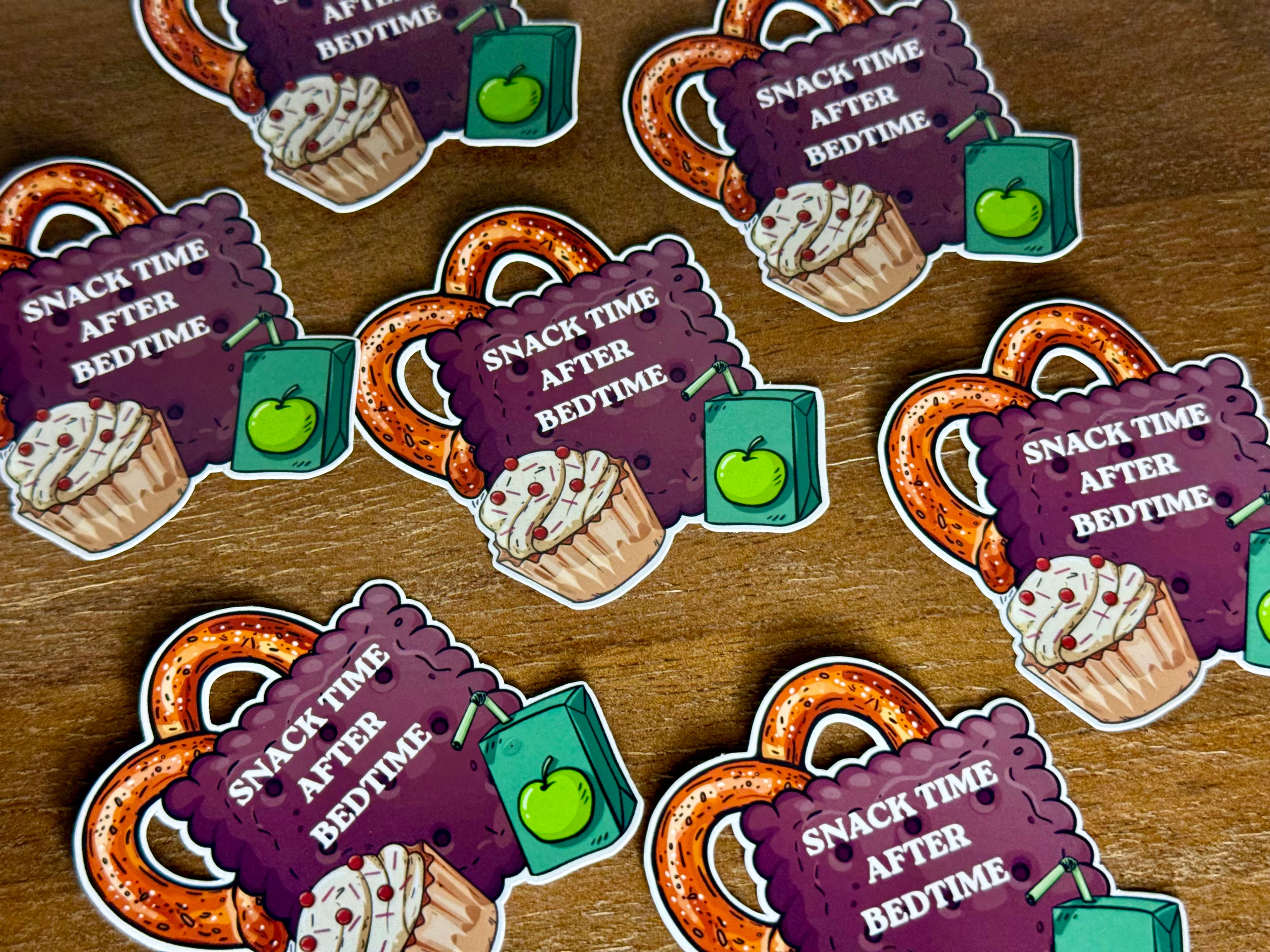 A set of die-cut stickers featuring a cartoon pretzel, a cupcake, a green apple, and a purple sign that reads 'SNACK TIME AFTER BEDTIME,' arranged on a wooden surface.