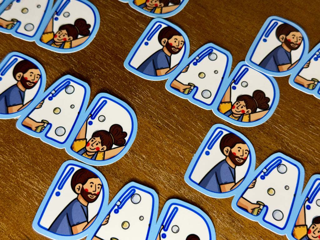 A close-up of multiple small die-cut stickers arranged on a wooden surface. Each sticker shows a cartoon illustration of a smiling man and girl in blue scrubs behind a bubble machine, with a blue border around the sticker.