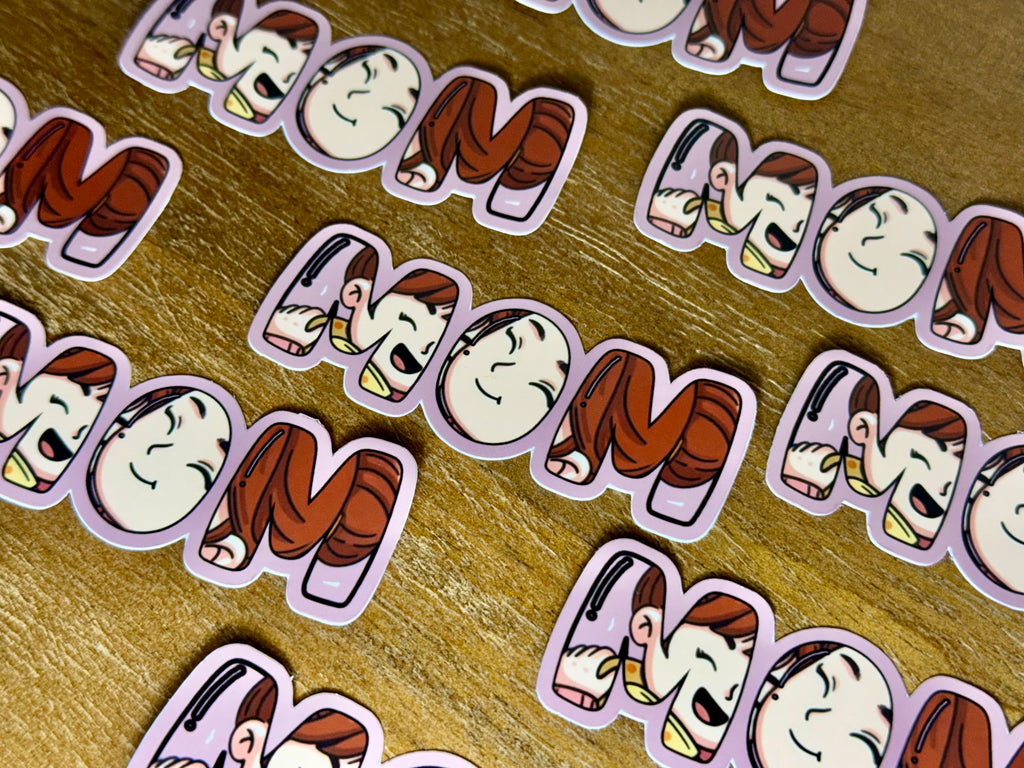 Multiple cartoon-style die-cut stickers spelling 'MOM' are arranged on a wooden surface. Each sticker features a smiling character with brown hair and a pink outfit inside a lavender outline.