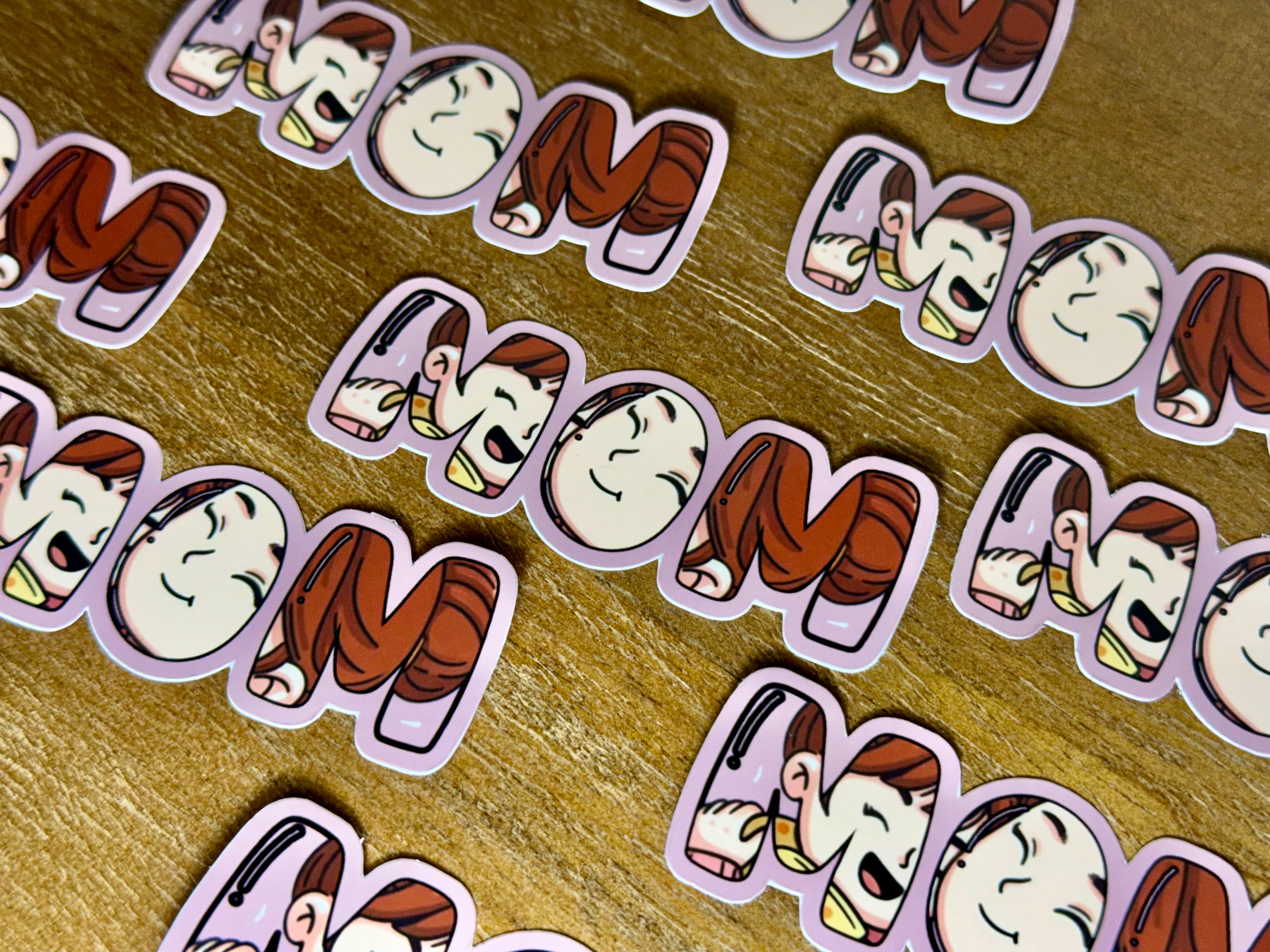 Multiple cartoon-style die-cut stickers spelling 'MOM' are arranged on a wooden surface. Each sticker features a smiling character with brown hair and a pink outfit inside a lavender outline.