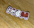 Cute "Mom" sticker featuring the word "Mom" with a loving mother and baby illustration incorporated into the lettering and finished with a transparent laminate.