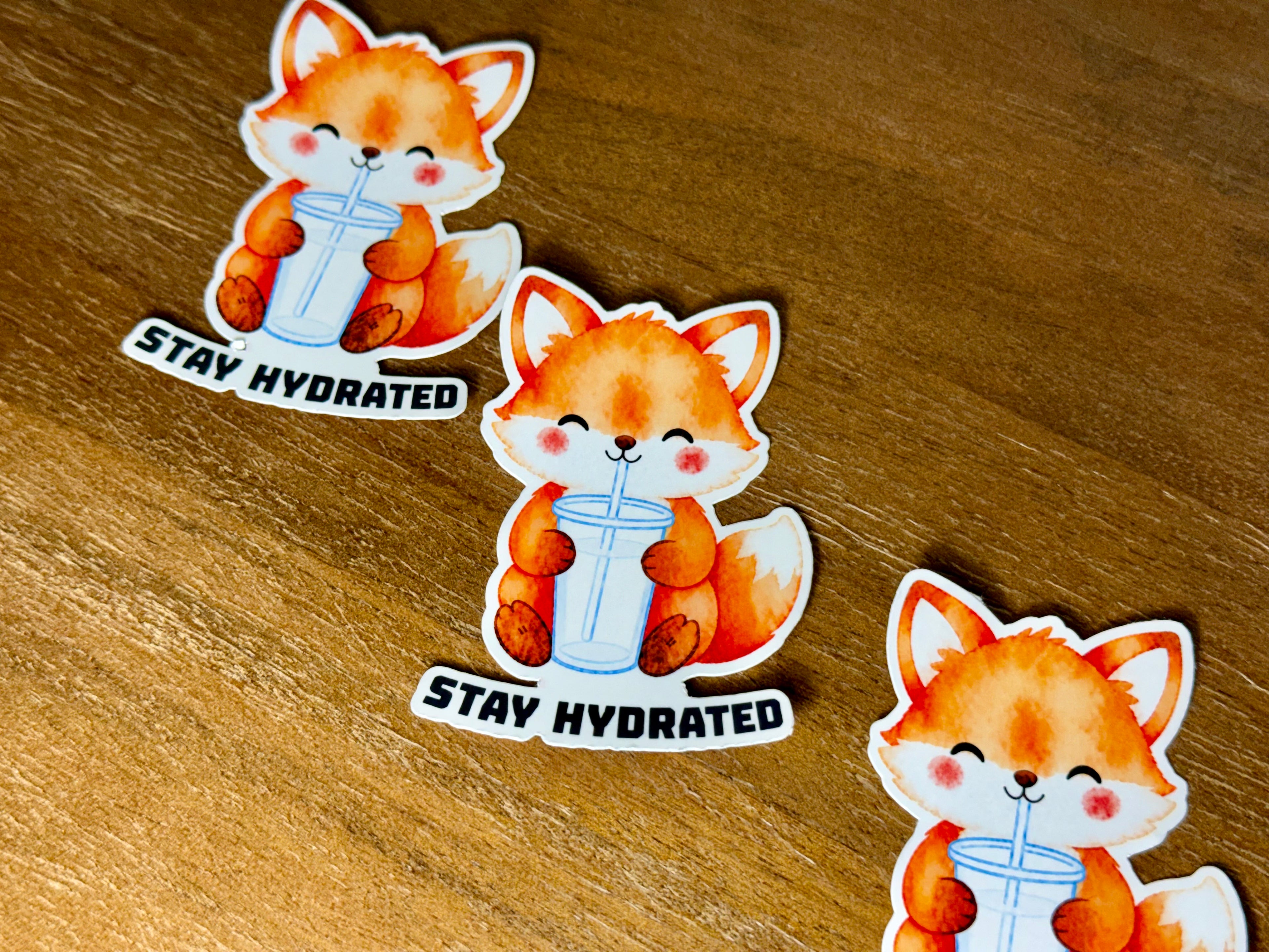 Three cartoon fox stickers drinking from a glass with the text 'STAY HYDRATED' arranged on a wooden surface.