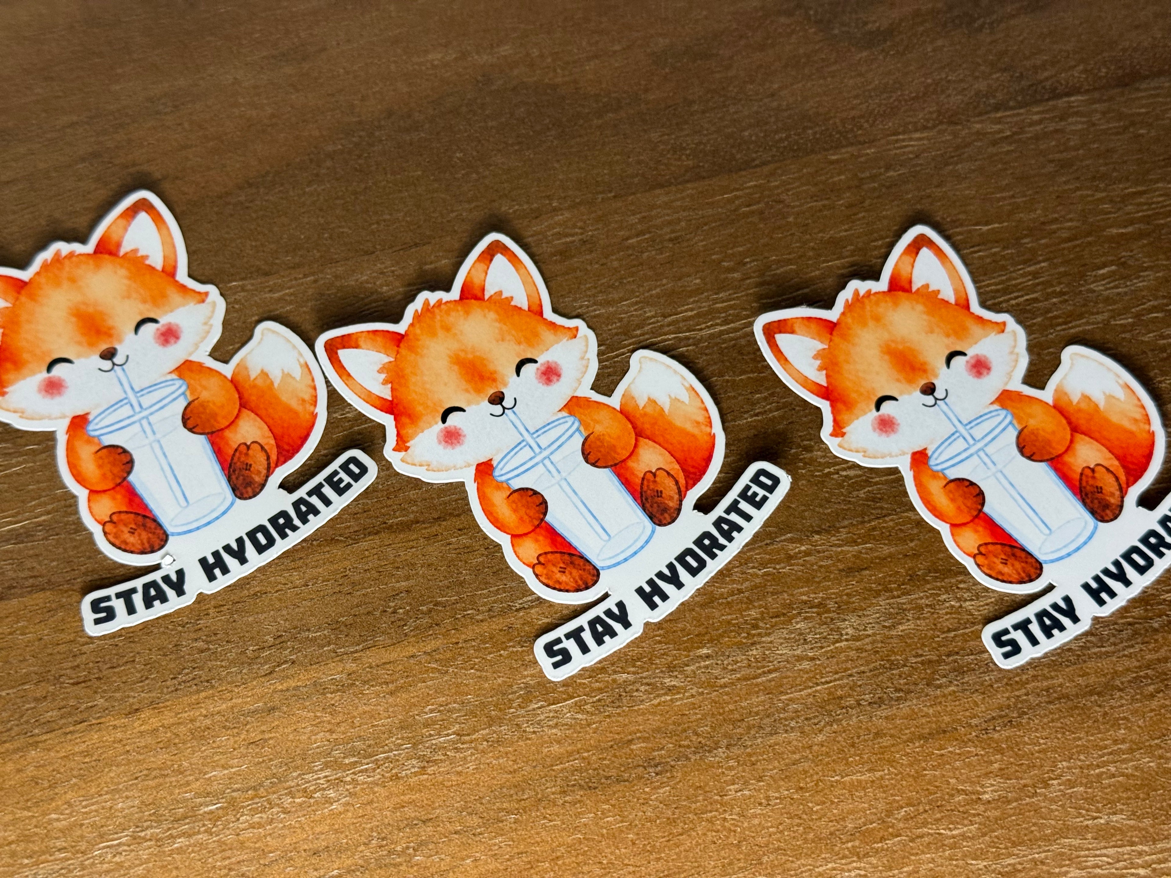Three identical die-cut stickers featuring a cute orange cartoon fox holding a white tumbler with the text 'STAY HYDRATED' underneath, arranged on a brown wooden surface.