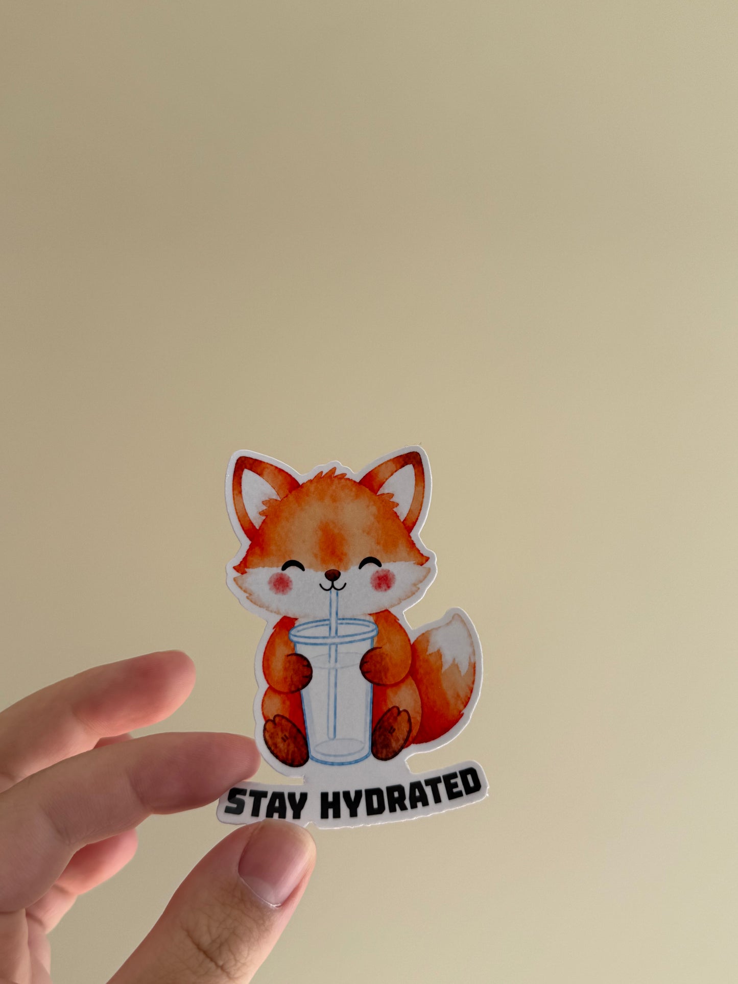 A hand holding a die-cut sticker featuring a cute orange fox drinking from a clear cup with the text 'STAY HYDRATED' underneath, set against a plain beige background.