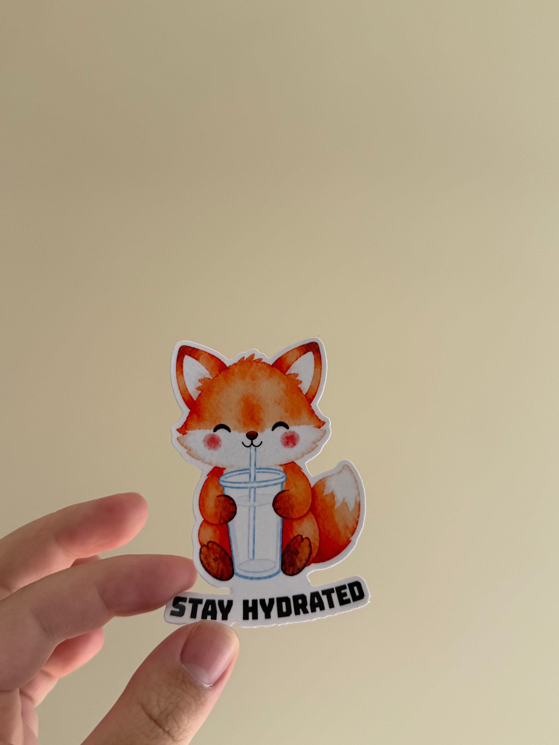 A hand holding a die-cut sticker featuring a cute orange fox drinking from a clear cup with the text 'STAY HYDRATED' underneath, set against a plain beige background.