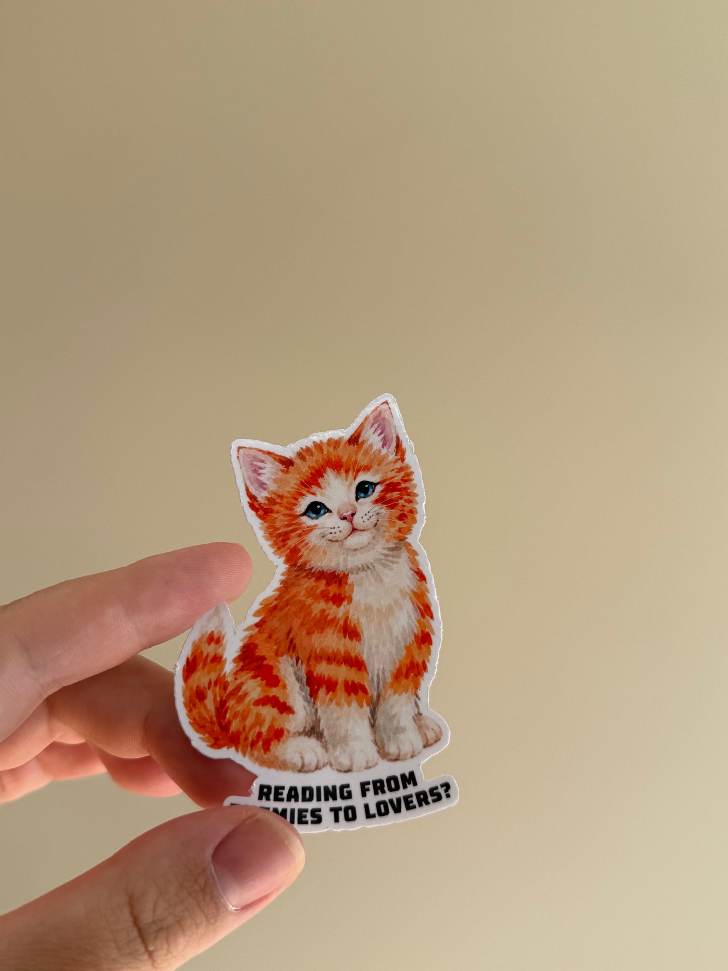 A hand holding a die-cut sticker of an orange tabby kitten with the text 'READING FROM EYES TO LOVERS?' against a plain beige background.