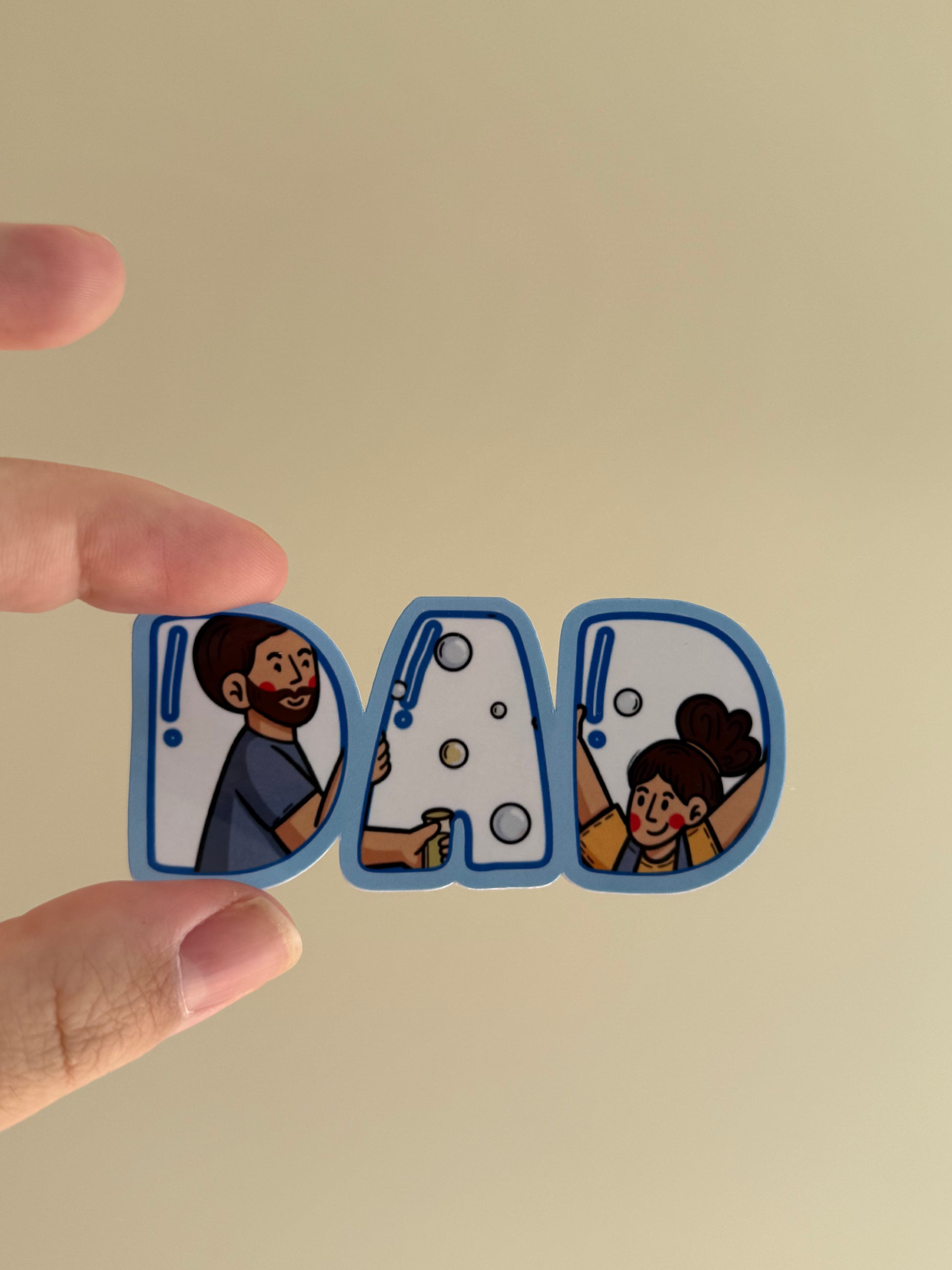 A close-up of a hand holding three die-cut letters spelling 'DAD' in a cartoon style. The letters are outlined in blue, with the left 'D' showing a man, the middle 'A' showing a child with bubbles, and the right 'D' showing a woman.