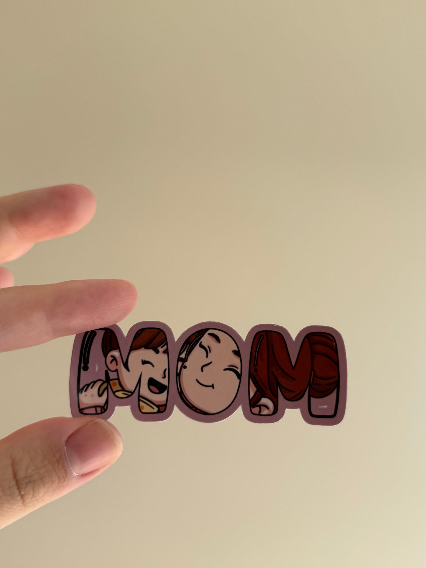 A hand holding a die-cut sticker spelling 'MOM' with cartoon character illustrations inside each letter, against a plain beige background.