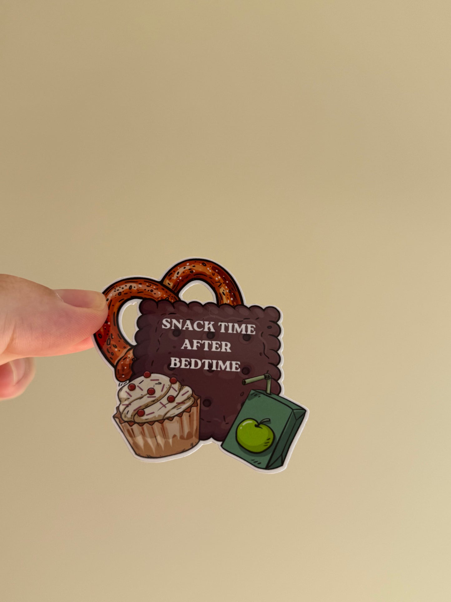 A hand holding a die-cut sticker featuring a pretzel, a cupcake, a green book with an apple, and a dark brown sign that reads 'SNACK TIME AFTER BEDTIME' against a plain beige background.