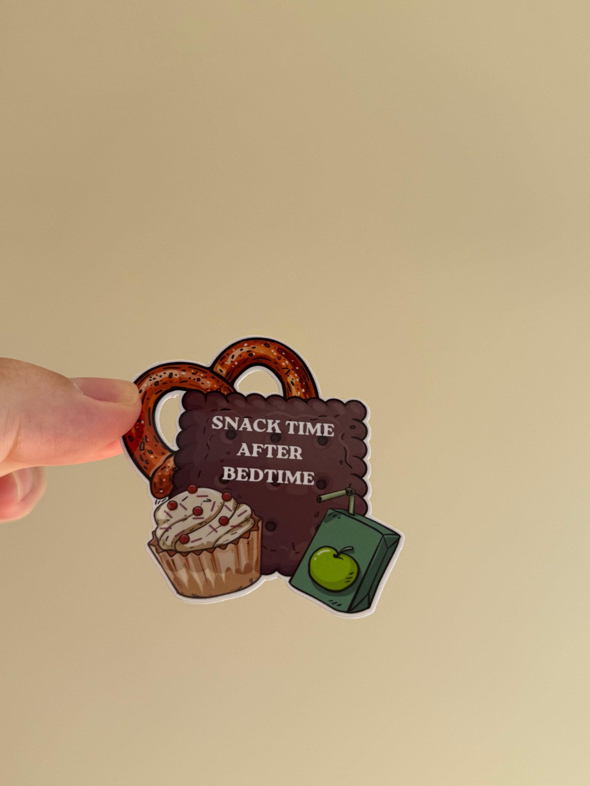 A hand holding a die-cut sticker featuring a pretzel, a cupcake, a green book with an apple, and a dark brown sign that reads 'SNACK TIME AFTER BEDTIME' against a plain beige background.