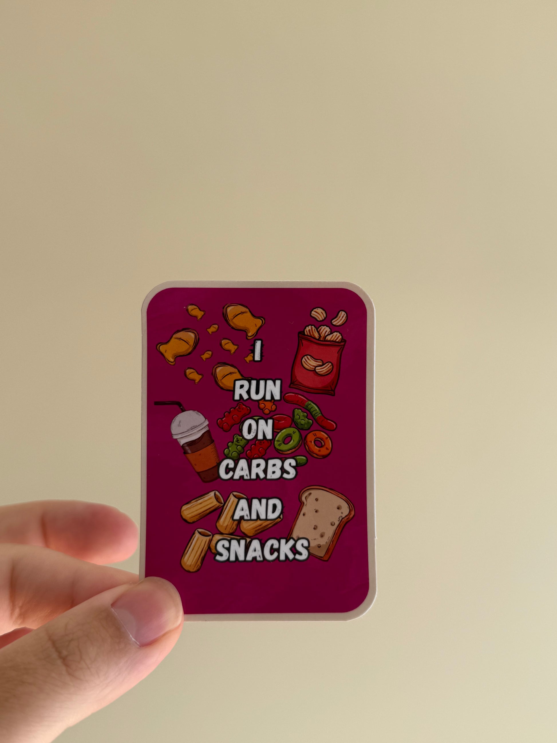 A hand holds a rectangular sticker with rounded corners against a plain beige background. The sticker has a dark magenta background with cartoon-style snack illustrations and the text 'I RUN ON CARBS AND SNACKS'.