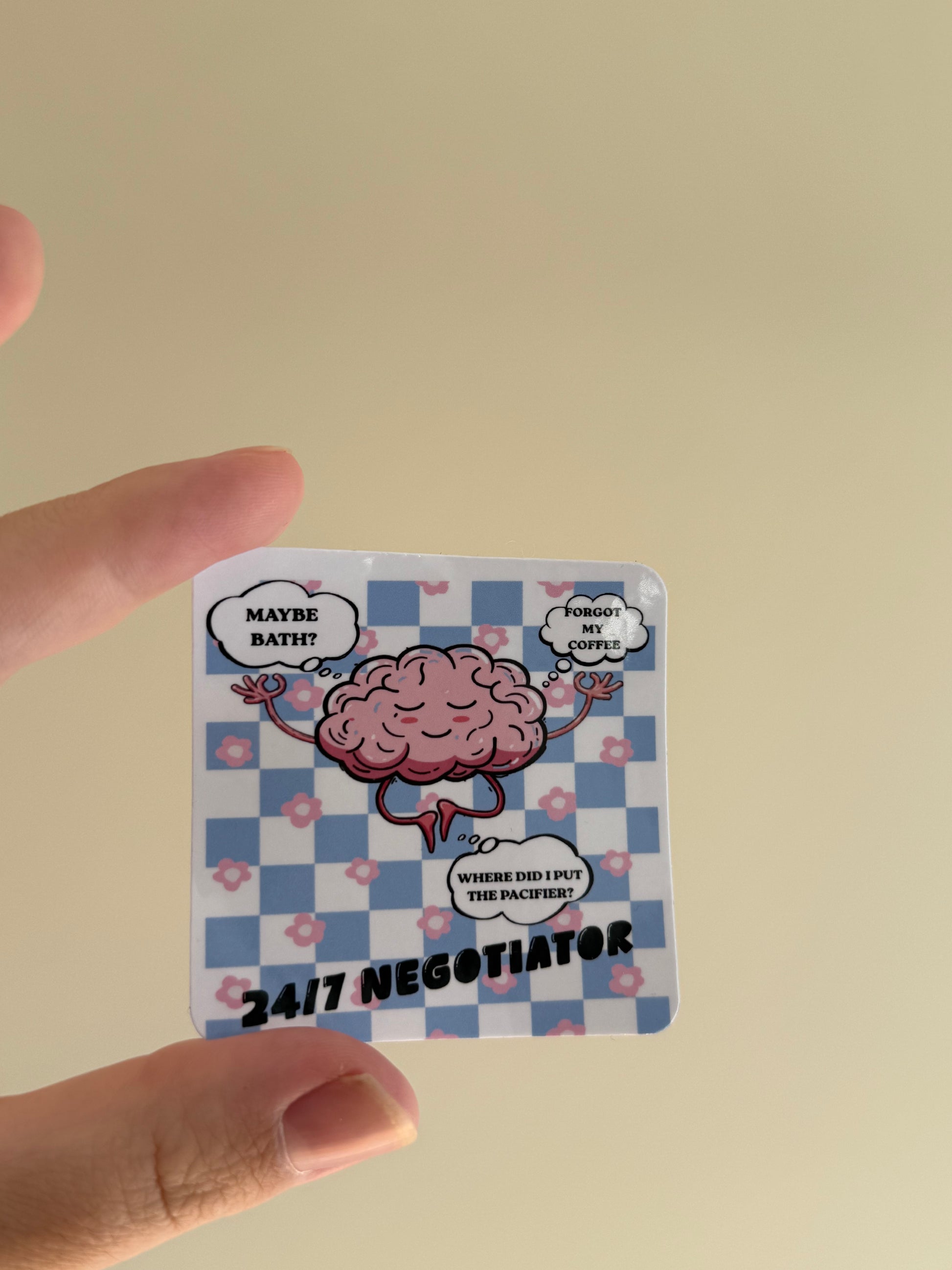 A hand holding a small square sticker or card featuring a cartoon pink brain on a blue-and-white checkered background with speech bubbles and the text '24/7 NEGOTIATOR'.