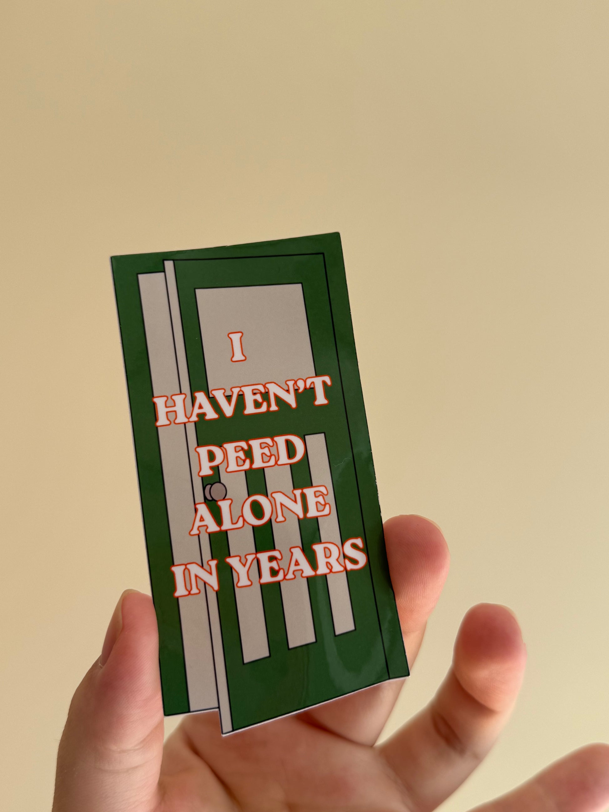 A hand holding a rectangular sticker featuring a green door graphic and the text 'I HAVEN'T PEED ALONE IN YEARS' in red letters, against a plain beige background.