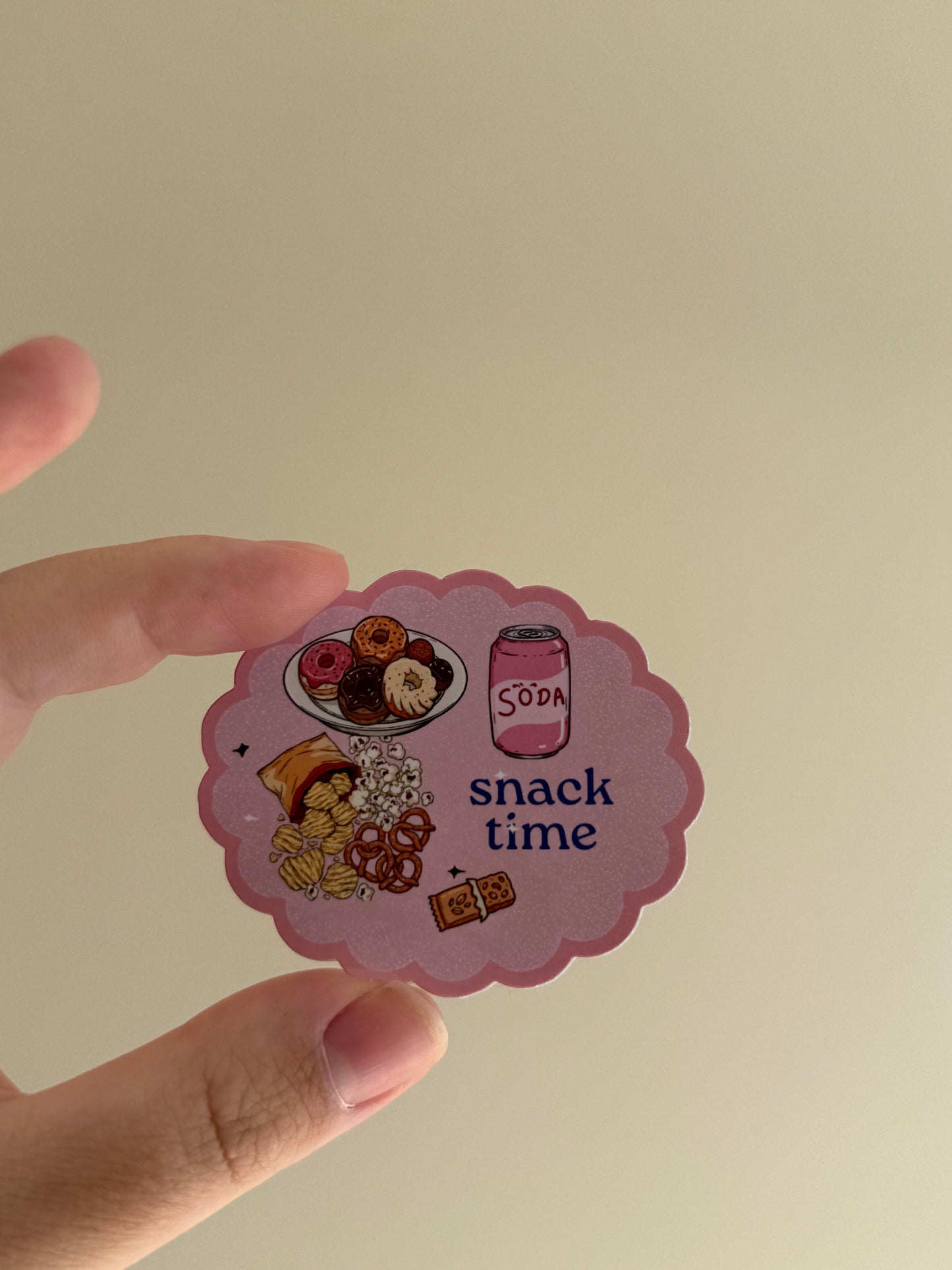 A hand holding a scalloped pink sticker that reads 'snack time' with illustrations of donuts, pretzels, candy, and a soda can, against a plain beige background.