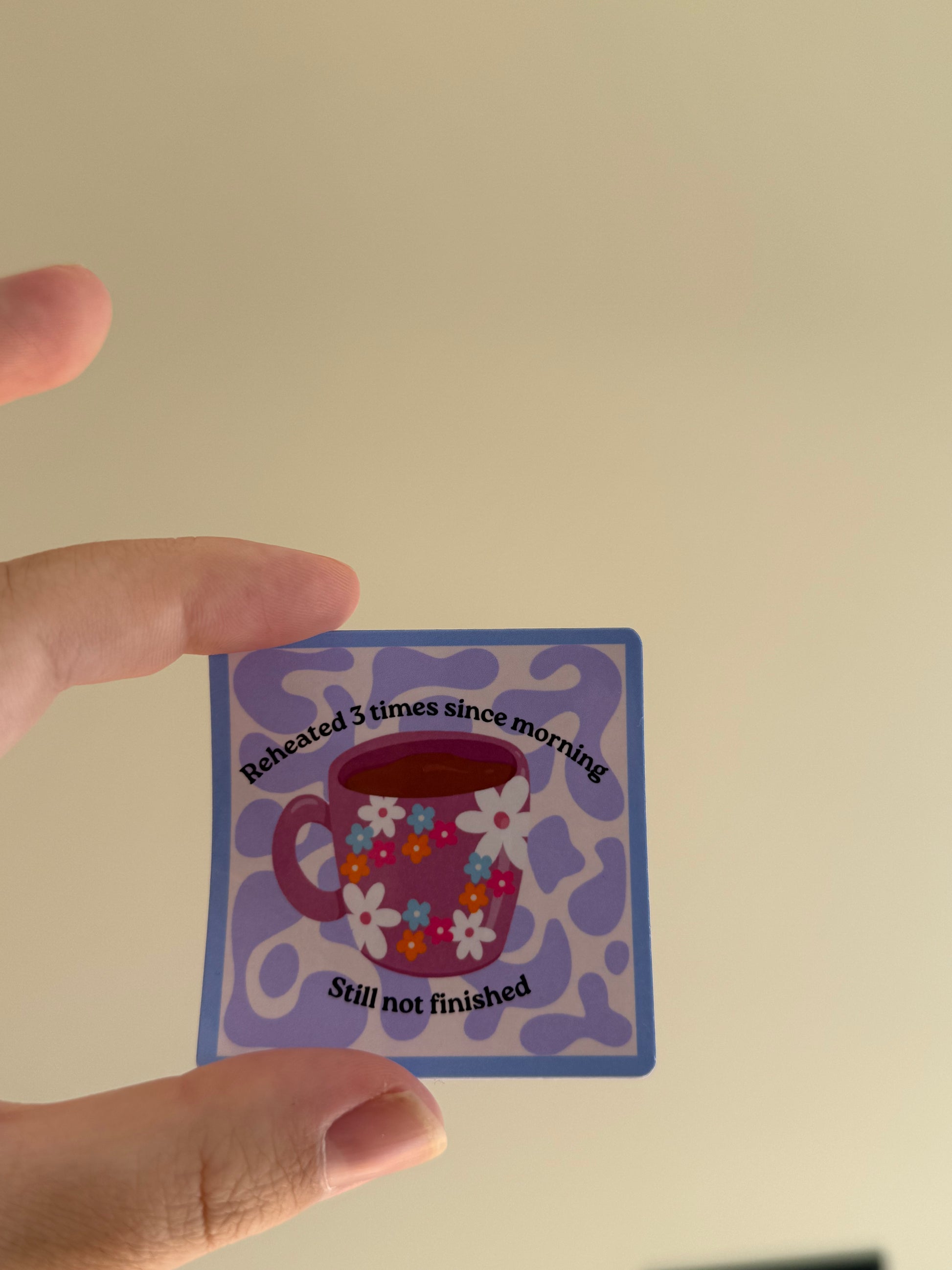 A hand holding a small square sticker with a purple coffee mug illustration and the text 'Reheated 3 times since morning' and 'Still not finished' against a plain beige wall.