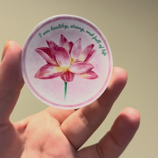 Close-up video showcasing a glitter laminate lotus affirmation sticker with shimmering finish and watercolor floral artwork from multiple angles.