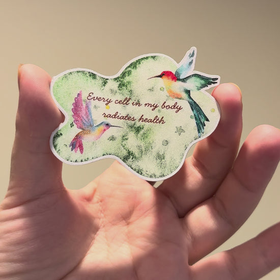 Close-up video of a watercolor hummingbird affirmation sticker with glitter laminate, showcasing its shimmering finish, vibrant colors, and handmade details from multiple angles.