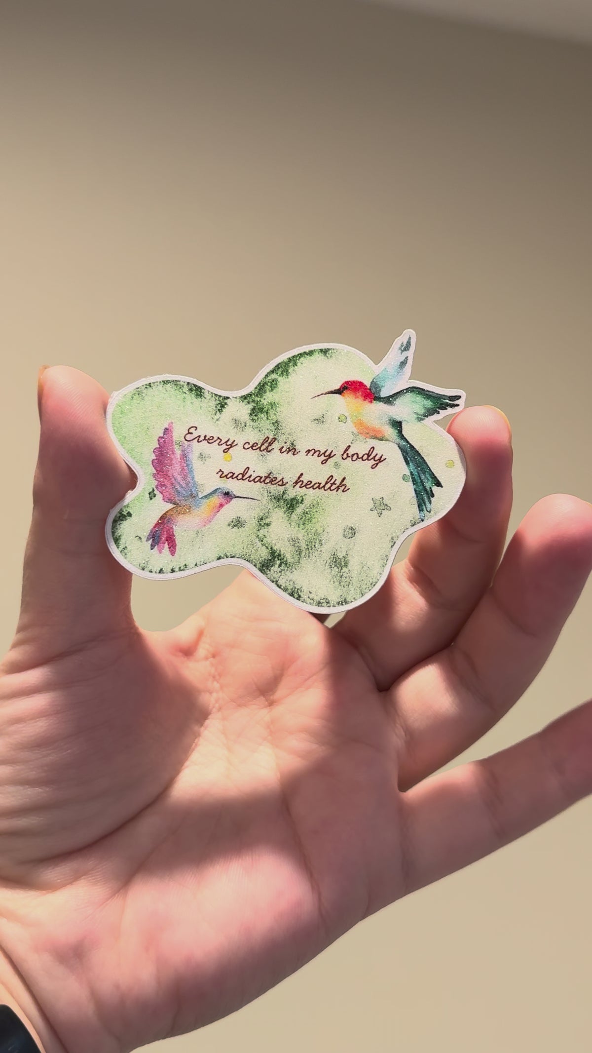 Close-up video of a watercolor hummingbird affirmation sticker with glitter laminate, showcasing its shimmering finish, vibrant colors, and handmade details from multiple angles.