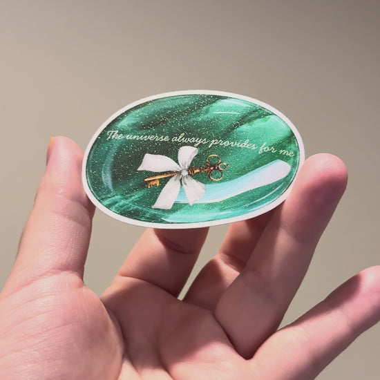 Hand holding a glitter affirmation sticker featuring an emerald crystal and the quote "The universe always provides for me.