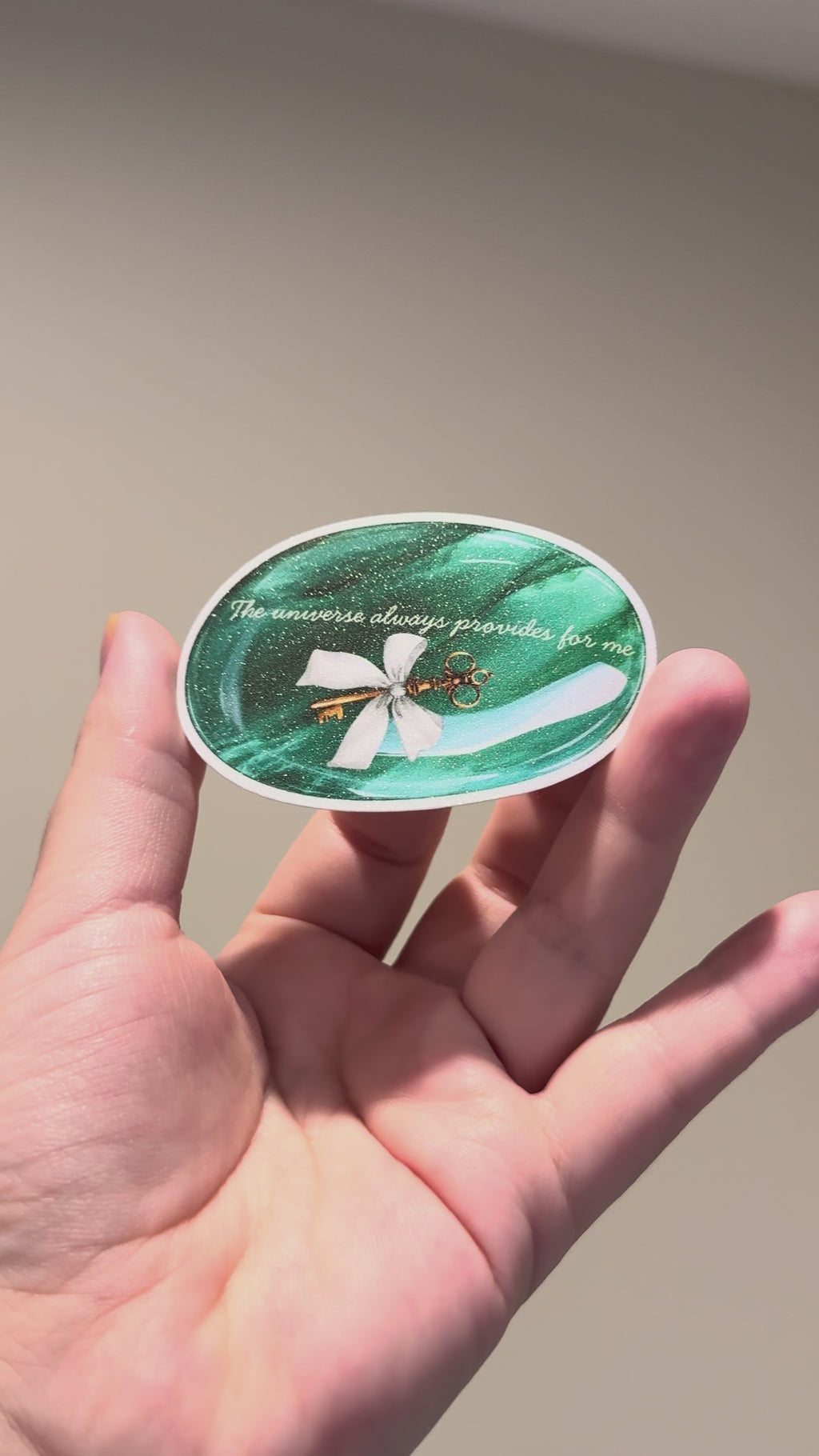 Hand holding a glitter affirmation sticker featuring an emerald crystal and the quote "The universe always provides for me.