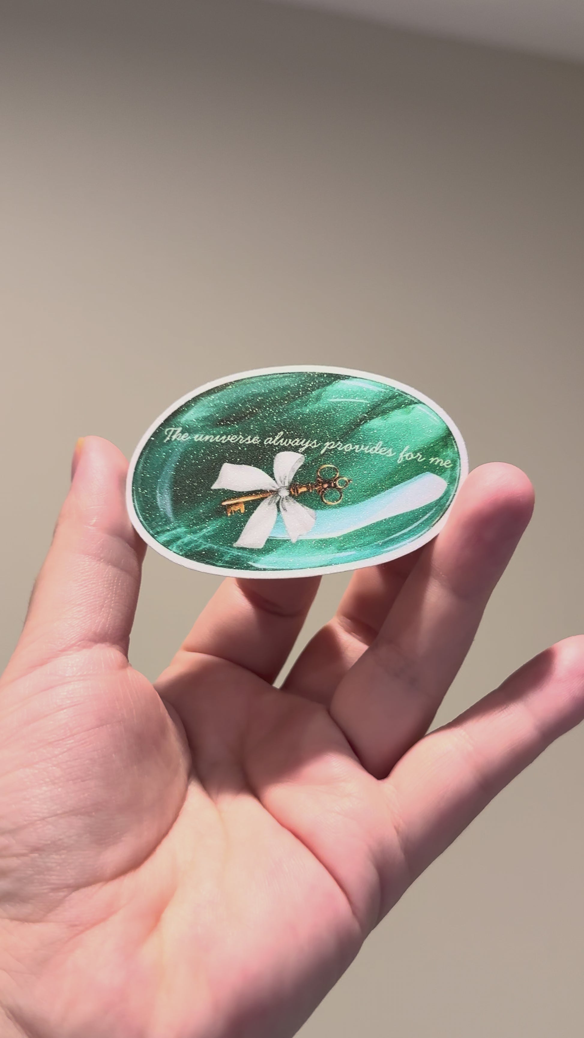 Hand holding a glitter affirmation sticker featuring an emerald crystal and the quote "The universe always provides for me.