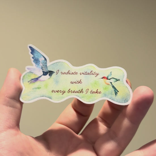 Close-up video of a watercolor bird affirmation sticker with glitter laminate, highlighting its shimmering finish, delicate artwork, and handmade details from multiple angles.