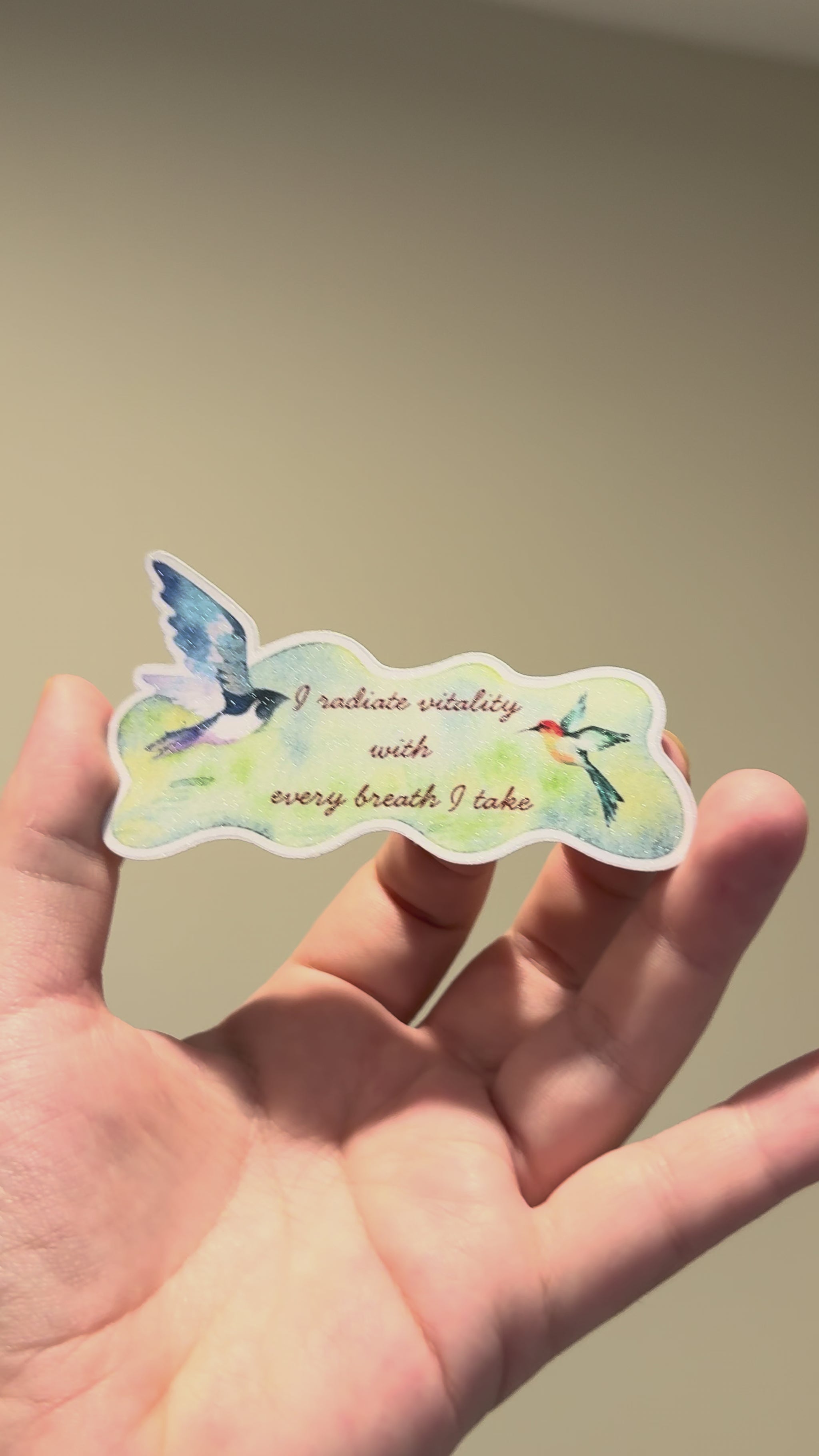 Close-up video of a watercolor bird affirmation sticker with glitter laminate, highlighting its shimmering finish, delicate artwork, and handmade details from multiple angles.